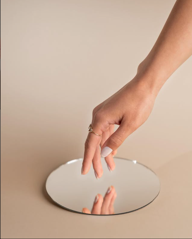 A manicured hand reaching toward a round mirror against a warm sand backdrop